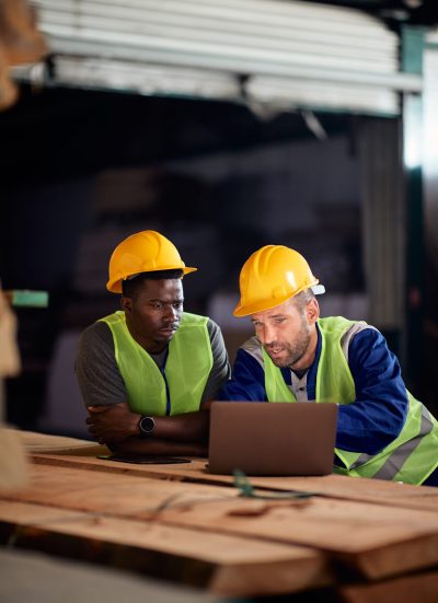 Warehouse workers cooperating while using laptop at wood department. Manual workers talking while working on a computer at lumber warehouse.